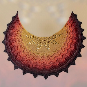 Puede incluir: Un chal de ganchillo en un degradado de colores que va del morado oscuro al naranja y al amarillo. El chal tiene forma de media luna y presenta un delicado patrón de encaje.