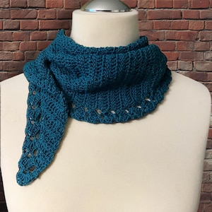 以下が含まれることがあります： マネキンに飾られた、ティール色の手編みスカーフ。 スカーフは、スカラップエッジと三角形の形状のテクスチャードデザインが特徴です。 背景はレンガの壁です。