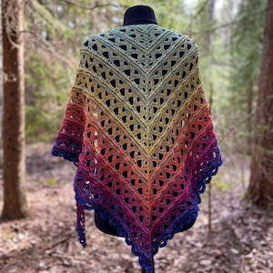 May include: A handmade, open-weave shawl in a gradient of colors, from green to yellow, orange, red, and finally, deep blue. The shawl is displayed on a black mannequin, showcasing its intricate crochet design and flowing shape. The background is a natural forest setting.