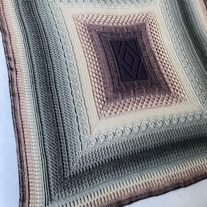 May include: A large, square crochet blanket with a multi-colored gradient pattern. The blanket is made with a variety of stitches and textures, creating a unique and intricate design. The colors range from light beige to dark purple, with shades of green and blue throughout.
