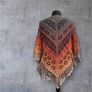 May include: A handcrafted crochet shawl in a gradient of colours, shifting from tan to orange and red. The shawl has a floral and geometric openwork design, displayed on a black mannequin head and stand against a grey background.