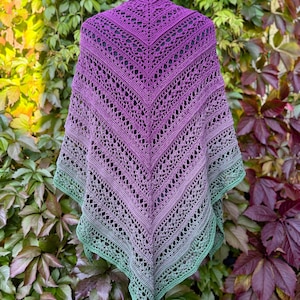 May include: A handmade, open-weave shawl in a gradient of colors, from deep purple at the top to a soft green at the bottom. The shawl is displayed on a black mannequin head against a backdrop of green and red foliage.