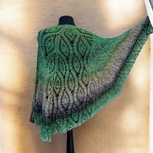 May include: A green and black crocheted shawl displayed on a mannequin. The shawl features a detailed openwork pattern with a gradient effect, transitioning from bright green to darker shades of green and black. The shawl is draped to showcase its design.
