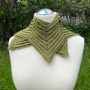 以下が含まれることがあります： 白いマネキンに飾られた、手作りのオリーブグリーンのクロシェ編みカウル。幾何学的なシェブロンデザインとスカラップエッジが特徴です。柔らかく、質感のある糸で作られており、暖かさとスタイルをプラスします。