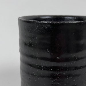 May include: A black ceramic tumbler with a textured, speckled surface. The cylindrical cup has a slightly flared rim and a dark, glossy finish. The handmade pottery piece is suitable for hot or cold beverages.