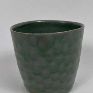 May include: A green ceramic cup with a textured surface. The cup is slightly wider at the top than the bottom.