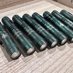 Op de afbeelding: Een verzameling donkergroene zaklampen met zilveren details. Elke zaklamp heeft een gegraveerde naam en nummer, zoals "Turslane Pack 66" en "July Pack". De zaklampen liggen op een houten oppervlak, wat hun ontwerp benadrukt.