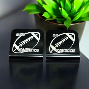 May include: Two black metal football-shaped desk organisers with white engraved footballs, numbers, and names. The organisers say "#95 TANNER" and "#2 TUCKER".