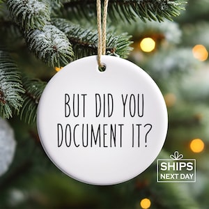 May include: A white ceramic Christmas ornament with the text "BUT DID YOU DOCUMENT IT?" in black. The ornament hangs from a gold string against a backdrop of a Christmas tree. The ornament also has the text "SHIPS NEXT DAY".