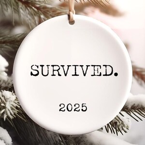 Puede incluir: Adorno navide&ntilde;o de cer&aacute;mica blanca con la palabra "SOBREVIV&Iacute;." en negro y el a&ntilde;o "2025". El adorno cuelga de una cuerda beige sobre un fondo de pino nevado.