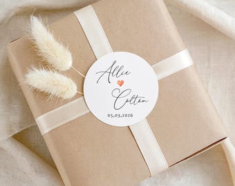 May include: A square gift box wrapped in brown paper with a cream-colored ribbon tied in a cross pattern. A round white sticker features the names Allie and Colton in black script, a small orange heart, and the date 05.03.2026. Two dried cream-colored bunny tail grasses are tucked under the ribbon.