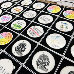 Puede incluir: Una colecci&oacute;n de adornos de cer&aacute;mica blanca con varios dise&ntilde;os y textos, incluyendo patrones tie-dye coloridos, gr&aacute;ficos tem&aacute;ticos familiares y citas inspiradoras. Cada adorno est&aacute; empaquetado en una caja negra.