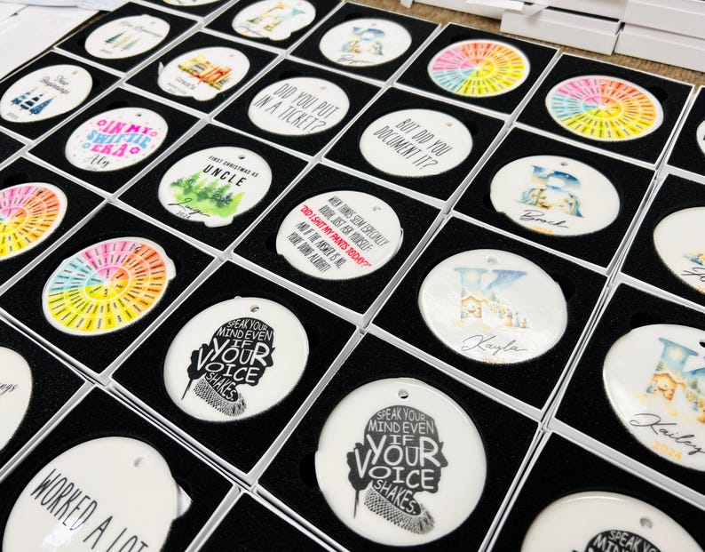 Puede incluir: Una colecci&oacute;n de adornos de cer&aacute;mica blanca con varios dise&ntilde;os y textos, incluyendo patrones tie-dye coloridos, gr&aacute;ficos tem&aacute;ticos familiares y citas inspiradoras. Cada adorno est&aacute; empaquetado en una caja negra.