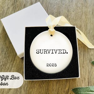 Puede incluir: Adorno de cer&aacute;mica blanca con la palabra "SURVIVED." y el a&ntilde;o "2025" impresos en negro. El adorno est&aacute; en una caja de regalo forrada en negro con una cinta crema. La caja es blanca.