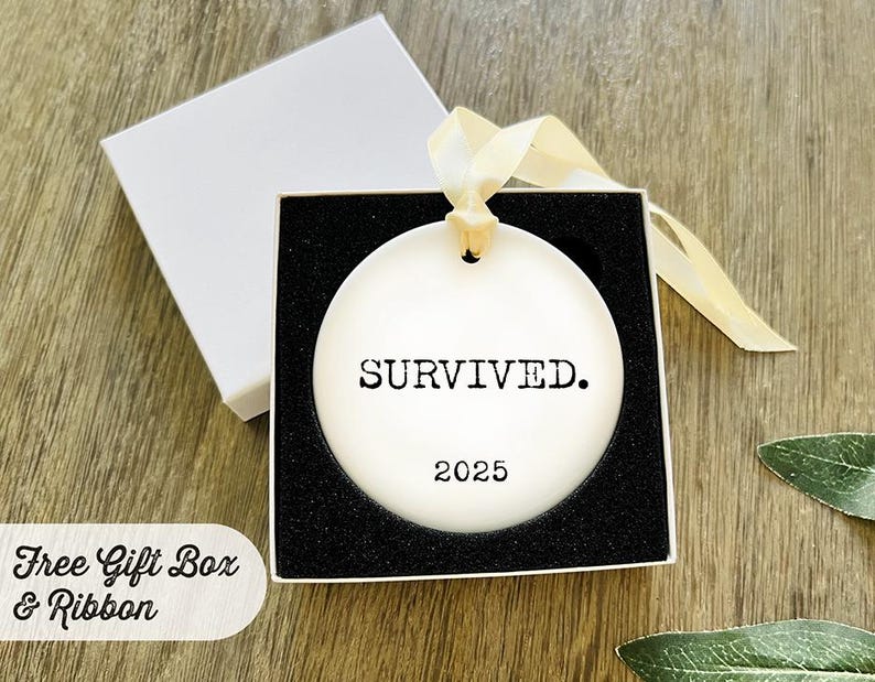 Puede incluir: Adorno de cer&aacute;mica blanca con la palabra "SURVIVED." y el a&ntilde;o "2025" impresos en negro. El adorno est&aacute; en una caja de regalo forrada en negro con una cinta crema. La caja es blanca.