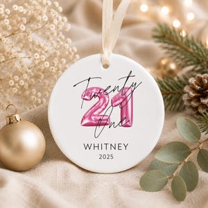May include: A round white ceramic ornament featuring a pink balloon-style number 21 graphic with the handwritten text Twenty One, the name WHITNEY, and the year 2025 printed in black. The ornament hangs from a light beige ribbon loop.