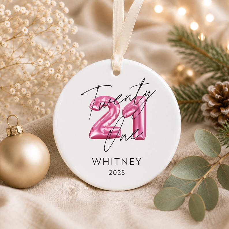 May include: A round white ceramic ornament featuring a pink balloon-style number 21 graphic with the handwritten text Twenty One, the name WHITNEY, and the year 2025 printed in black. The ornament hangs from a light beige ribbon loop.