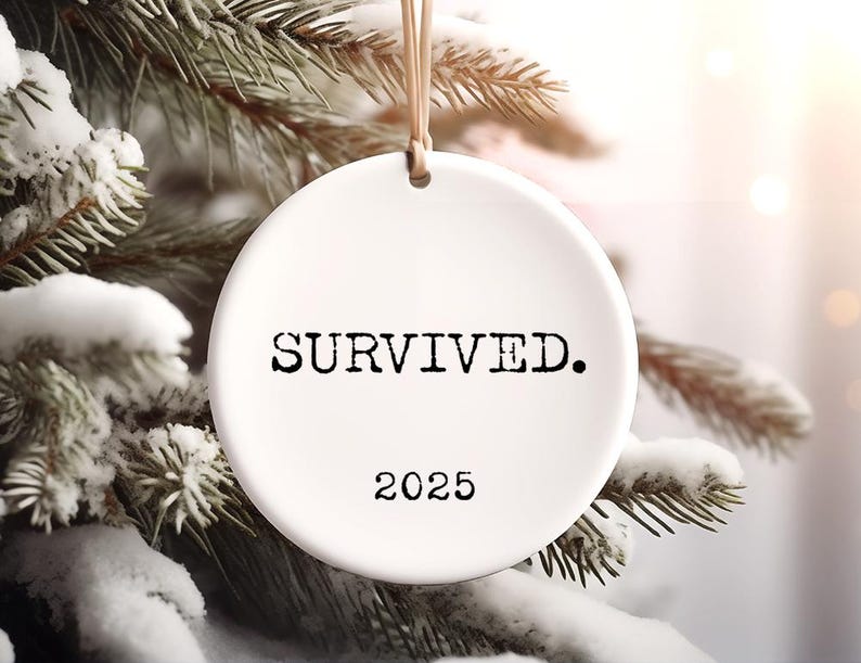 Puede incluir: Adorno navide&ntilde;o de cer&aacute;mica blanca con la palabra "SOBREVIV&Iacute;." en negro y el a&ntilde;o "2025". El adorno cuelga de una cuerda beige sobre un fondo de pino nevado.