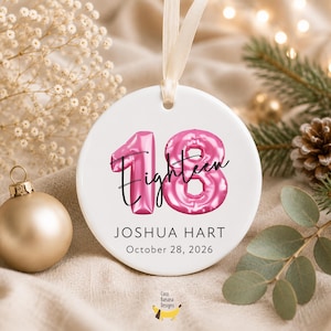 May include: A round white ceramic ornament hanging from a cream ribbon. The design features large pink metallic balloon-style numbers 18, the word Eighteen in black script, and the text JOSHUA HART and October 28, 2026, printed in black.