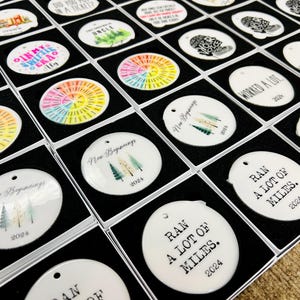 Puede incluir: Una colecci&oacute;n de adornos blancos y redondos en cajas negras. Algunos adornos tienen dise&ntilde;os coloridos, mientras que otros presentan texto como "New Beginnings 2024" y "Ran a lot of miles 2024". Los adornos est&aacute;n listos para exhibir.