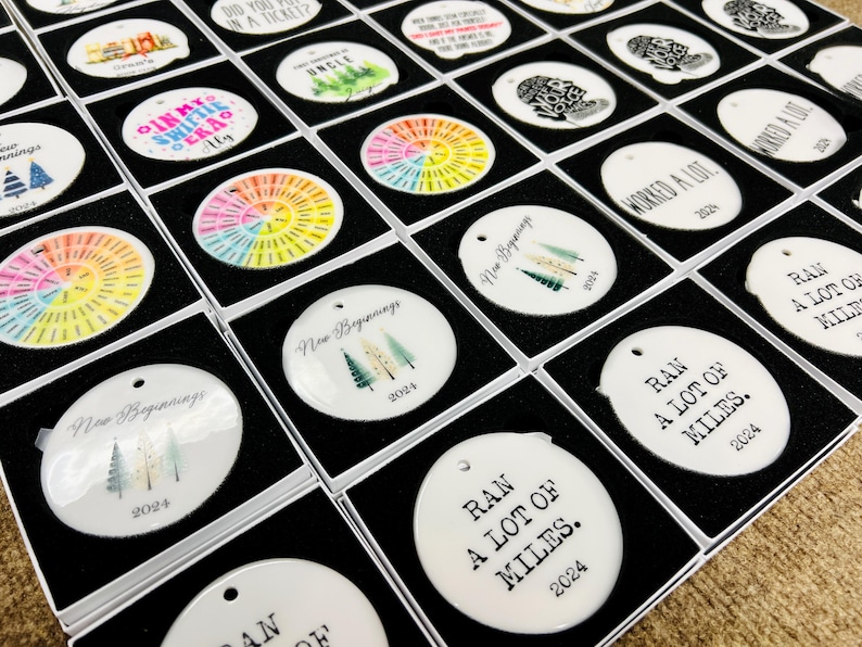 Puede incluir: Una colecci&oacute;n de adornos blancos y redondos en cajas negras. Algunos adornos tienen dise&ntilde;os coloridos, mientras que otros presentan texto como "New Beginnings 2024" y "Ran a lot of miles 2024". Los adornos est&aacute;n listos para exhibir.