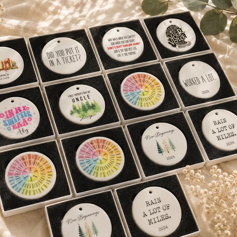 May include: A collection of round white ceramic ornaments displayed in individual black-lined gift boxes. The ornaments feature various designs including colourful radial charts, evergreen tree illustrations, and text such as "Did you put it in a ticket?", "First Father's Day as Uncle", "Worked a lot 2024", and "Rain a lot of miles 2024".