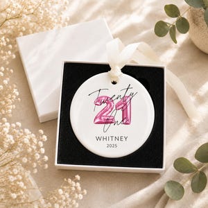 May include: A round white ceramic ornament featuring a pink balloon-style number 21 graphic with the handwritten text Twenty One above the name WHITNEY and the year 2025 in black font, tied with a cream ribbon and presented in a black-lined gift box.
