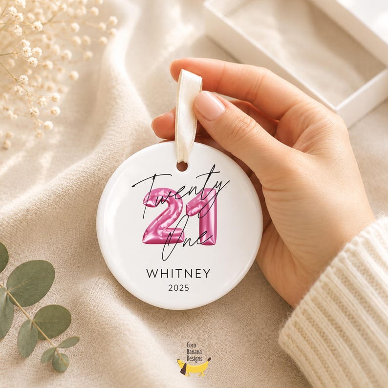 May include: A round white ceramic ornament featuring a pink metallic balloon-style number 21 graphic, the handwritten script text Twenty One, the name WHITNEY, and the year 2025 in black print, suspended by a white satin ribbon loop.