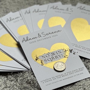 May include: A stack of grey cards with gold foil hearts. The cards have the text "Adam & Serena" and "22nd December 2024" printed on them. The top card has a gold foil heart with a scratch-off area that says "No Win Sorry!" and a coin underneath.