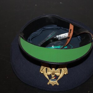 以下が含まれることがあります： 金色のエンブレムと緑色の内装が付いた、濃紺のミリタリーベレー帽。ベレー帽には黒い革バンドと茶色の革ストラップが付いています。エンブレムは、青と赤のアクセントが付いた金のワシです。