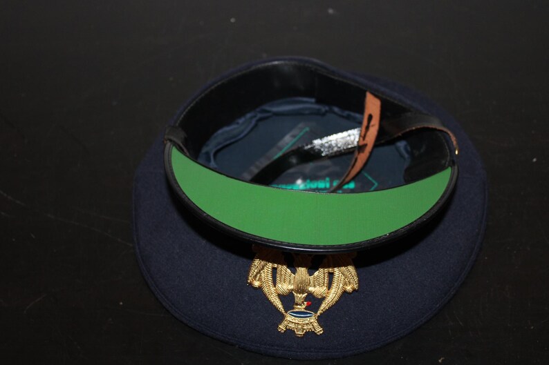 以下が含まれることがあります： 金色のエンブレムと緑色の内装が付いた、濃紺のミリタリーベレー帽。ベレー帽には黒い革バンドと茶色の革ストラップが付いています。エンブレムは、青と赤のアクセントが付いた金のワシです。