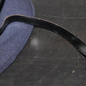 以下が含まれることがあります： 黒い革ストラップが付いた濃紺の帽子のクローズアップ。ストラップは金色のボタンで帽子に取り付けられています。帽子には緑色の裏地が付いています。