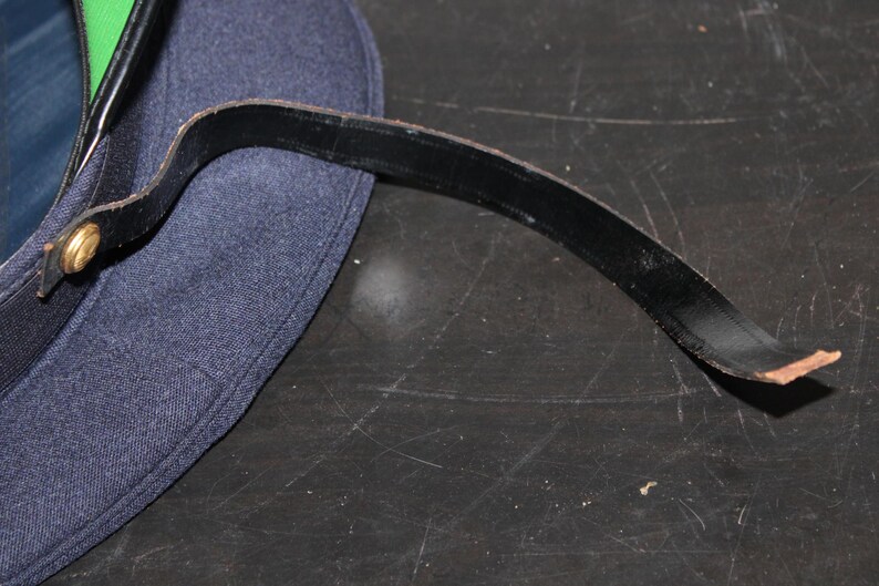 以下が含まれることがあります： 黒い革ストラップが付いた濃紺の帽子のクローズアップ。ストラップは金色のボタンで帽子に取り付けられています。帽子には緑色の裏地が付いています。