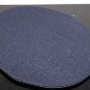 以下が含まれることがあります： 平らなトップが付いた、ネイビーブルーの円形帽子。帽子はテクスチャード加工された生地で作られており、制服の帽子のように見えます。帽子は単色で、シンプルなデザインです。