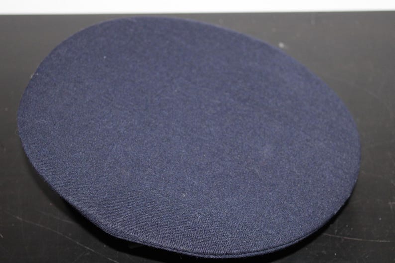 以下が含まれることがあります： 平らなトップが付いた、ネイビーブルーの円形帽子。帽子はテクスチャード加工された生地で作られており、制服の帽子のように見えます。帽子は単色で、シンプルなデザインです。