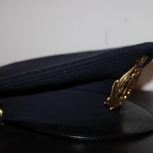 以下が含まれることがあります： 黒いひさしと金色のエンブレムが付いた濃いネイビーブルーの帽子。帽子は丸みを帯びたクラウンと側面にボタンが付いています。エンブレムは、様式化された葉または翼のデザインです。