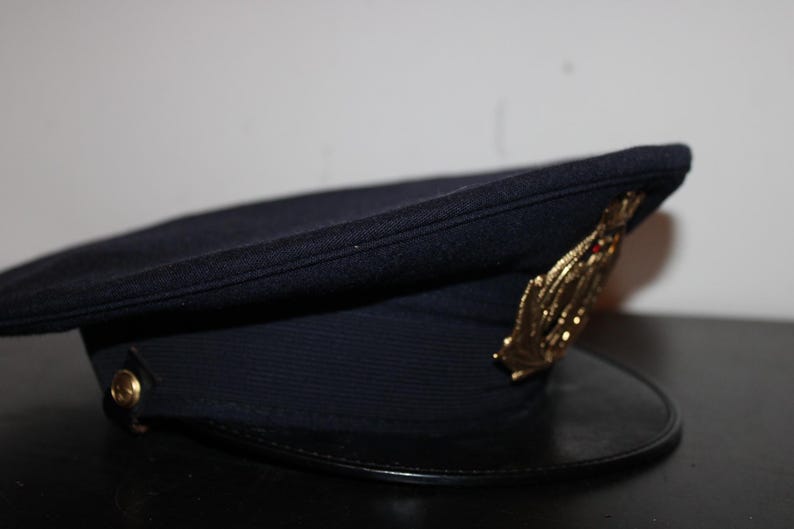 以下が含まれることがあります： 黒いひさしと金色のエンブレムが付いた濃いネイビーブルーの帽子。帽子は丸みを帯びたクラウンと側面にボタンが付いています。エンブレムは、様式化された葉または翼のデザインです。