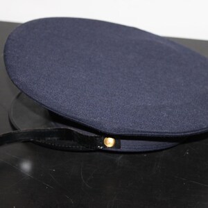 以下が含まれることがあります： 丸みを帯びたクラウンと黒いブリムが付いた濃いネイビーブルーの帽子。帽子には金色のボタンと黒いストラップが付いています。帽子は暗い表面に置かれています。