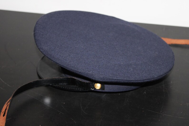 以下が含まれることがあります： 丸みを帯びたクラウンと黒いブリムが付いた濃いネイビーブルーの帽子。帽子には金色のボタンと黒いストラップが付いています。帽子は暗い表面に置かれています。