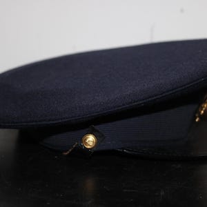 以下が含まれることがあります： 丸みを帯びたトップと黒いツバが付いた濃紺の帽子。 金色のエンブレムとボタンのディテールが特徴です。 この帽子はおそらく制服の帽子です。