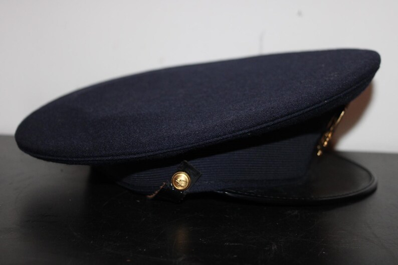 以下が含まれることがあります： 丸みを帯びたトップと黒いツバが付いた濃紺の帽子。 金色のエンブレムとボタンのディテールが特徴です。 この帽子はおそらく制服の帽子です。