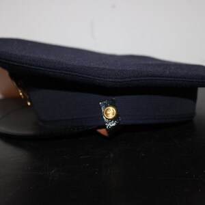 以下が含まれることがあります： 黒い縁が付いた濃い紺色の帽子。帽子には金色のボタンと金色のエンブレムが付いています。帽子は黒い表面に置かれています。