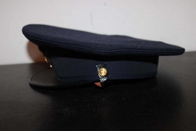 以下が含まれることがあります： 黒い縁が付いた濃い紺色の帽子。帽子には金色のボタンと金色のエンブレムが付いています。帽子は黒い表面に置かれています。