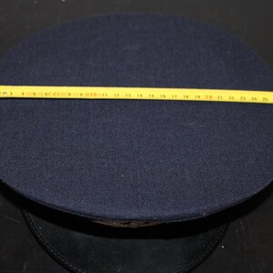 以下が含まれることがあります： 円形のクラウンが付いた濃紺の帽子。黄色の巻尺が帽子の直径を示しています。帽子はフェルトのような素材でできているようです。帽子は黒い表面に置かれています。