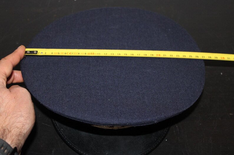 以下が含まれることがあります： 円形のクラウンが付いた濃紺の帽子。黄色の巻尺が帽子の直径を示しています。帽子はフェルトのような素材でできているようです。帽子は黒い表面に置かれています。