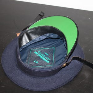 以下が含まれることがあります： グリーンのつばと黒革のトリムが付いたネイビーブルーの帽子。内側はネイビーブルーで、ラベルが付いています。黒革のストラップが帽子に付いています。サイズ57。