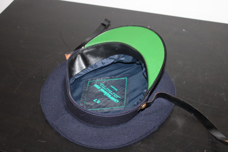 以下が含まれることがあります： グリーンのつばと黒革のトリムが付いたネイビーブルーの帽子。内側はネイビーブルーで、ラベルが付いています。黒革のストラップが帽子に付いています。サイズ57。