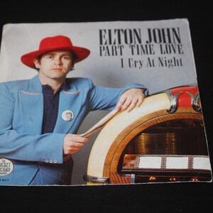 Könnte beinhalten: Eine Vintage-Schallplattenhülle mit Elton John in einem blauen Anzug und rotem Hut, der sich an einen Jukebox lehnt. Der Text auf der Hülle lautet "Elton John, Part Time Love, I Cry At Night".