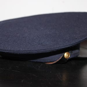 以下が含まれることがあります： 丸みを帯びたトップと黒いブリムが付いた濃いネイビーブルーの帽子。 金色のボタンが側面にあります。 帽子は、テクスチャード加工された生地で作られているようです。