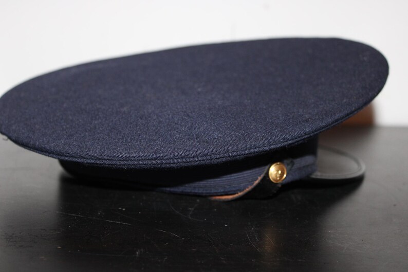 以下が含まれることがあります： 丸みを帯びたトップと黒いブリムが付いた濃いネイビーブルーの帽子。 金色のボタンが側面にあります。 帽子は、テクスチャード加工された生地で作られているようです。